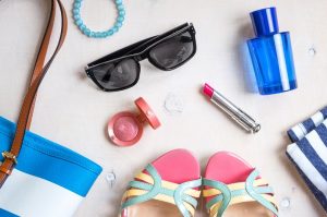 Set of summer women's accessories: sunglasses, shoes, slippers, passport, blue striped bag, pink lipstick, blush, perfume on white wood background. Feminine vacation/travel/sea objects. Top view